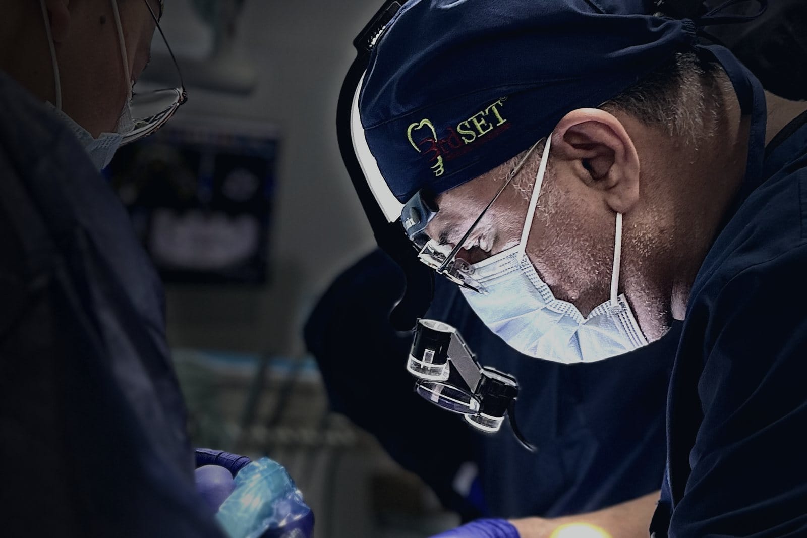 A masked clinician wearing loupes and a 3rdSET surgical cap works close to a patient's mouth during live-patient training
